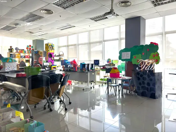Bright classroom with colorful children’s crafts, small tables, and chairs near large windows.