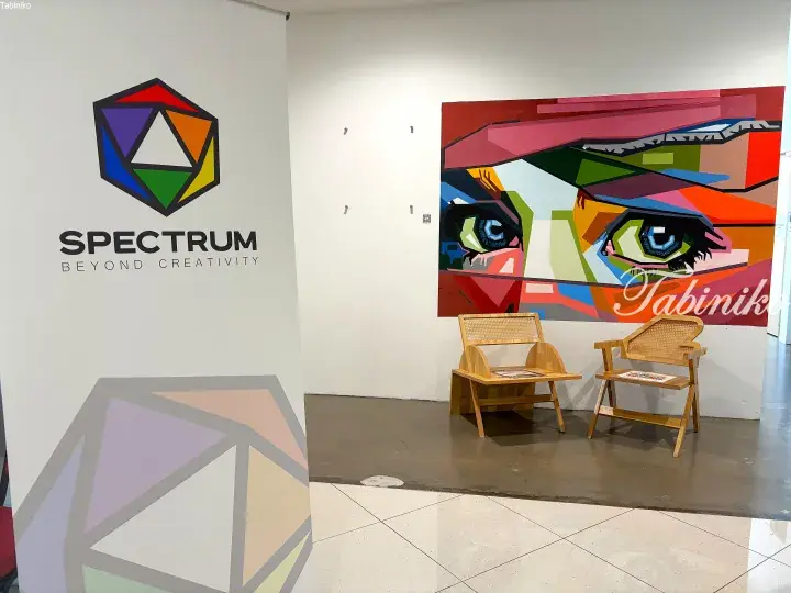 Spectrum Beyond Creativity logo and colorful abstract eye artwork displayed in a modern gallery space with wooden chairs.