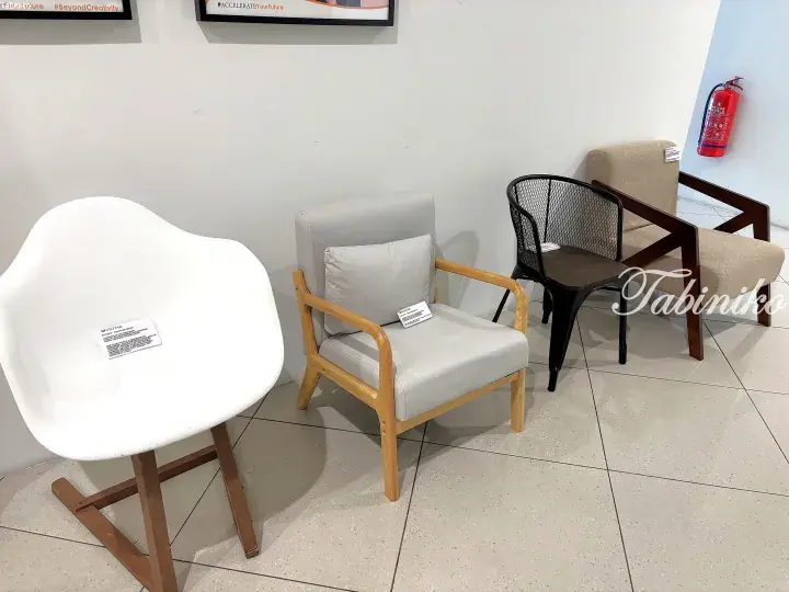 A display of modern furniture including a white molded chair, a light wood armchair with cushions, and a black wire chair in a showroom.