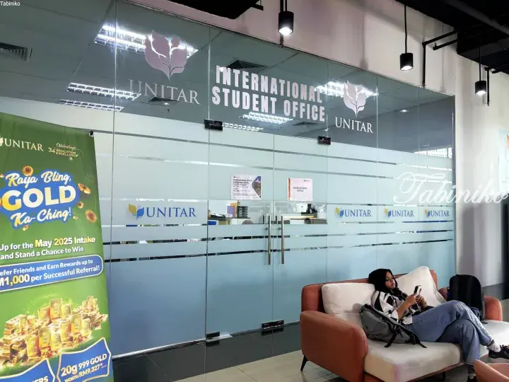 A student relaxes on a sofa outside UNITAR’s International Student Office, with a Raya gold promotion banner visible nearby.