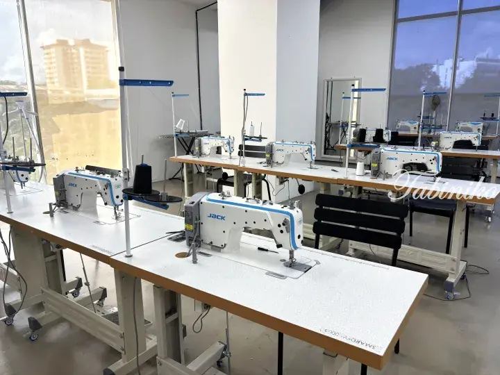 Rows of industrial sewing machines on worktables in a bright, modern garment workshop with large windows.