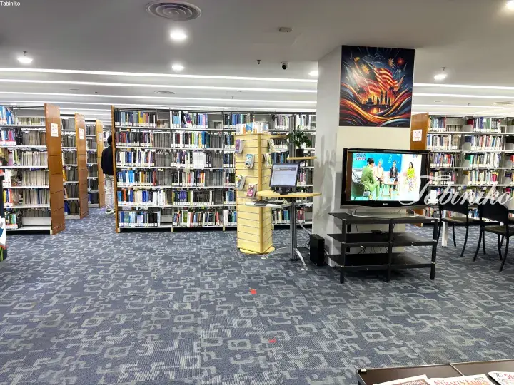 Modern library interior with bookshelves, a TV showing a Malaysian talk show, and a patriotic wall mural.