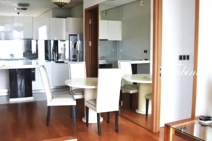 Modern dining area with white chairs and a round table, featuring a sleek kitchen and large mirror in a Malaysian apartment.