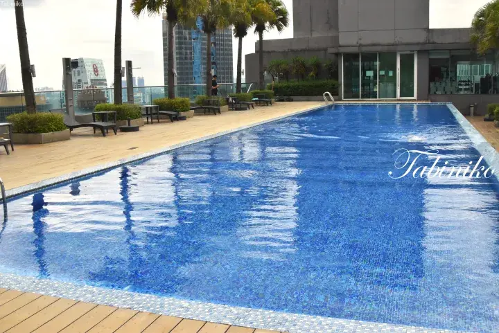 Rooftop swimming pool with wooden deck and palm trees overlooking Kuala Lumpur city skyline.