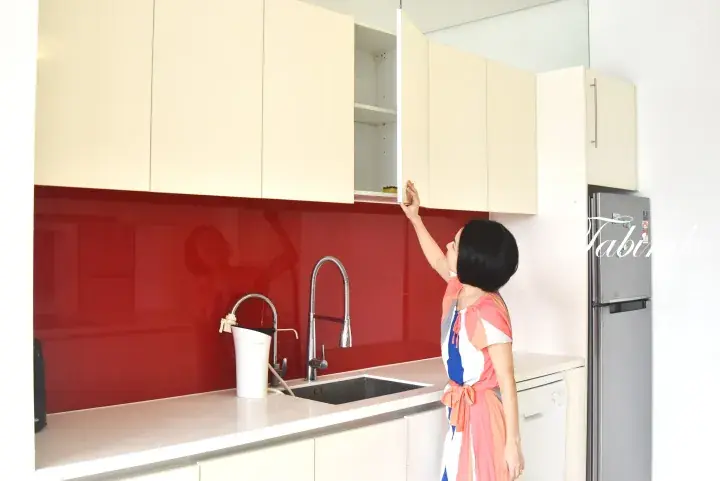 Woman opening a cream kitchen cabinet above a red backsplash in a modern apartment kitchen.