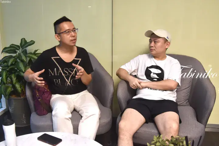 Two men conversing in a modern lounge, one wearing a black ‘MASSIVE BLACK’ t-shirt and the other a white BAPE shirt.