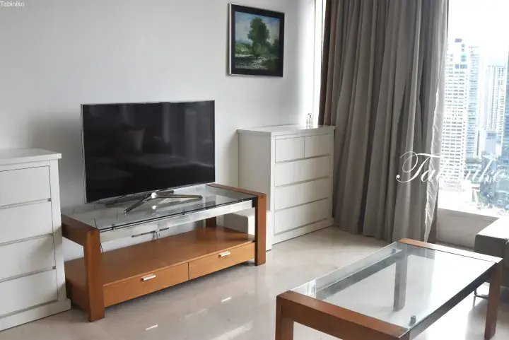 Modern living room with a flat-screen TV on a wooden stand, white drawers, and a glass coffee table overlooking a city skyline.