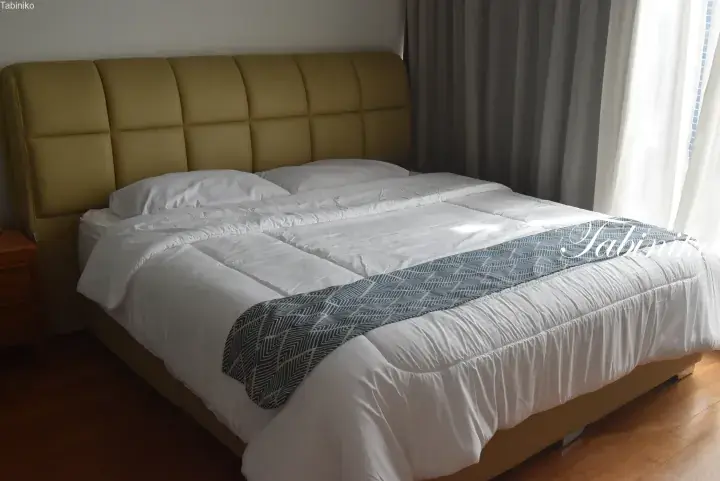 A comfortable bed with a mustard-yellow tufted headboard, white linens, and a blue patterned runner in a bright room.