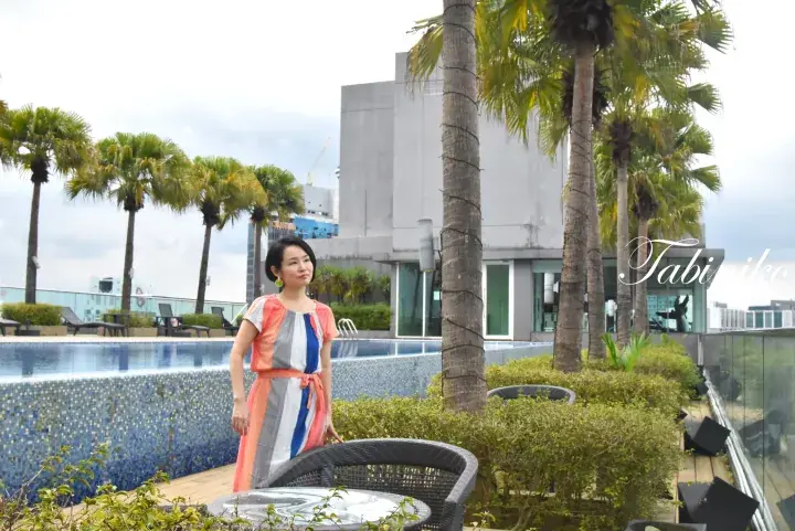 Woman in a colorful dress standing by a rooftop pool with palm trees and modern buildings in the background.