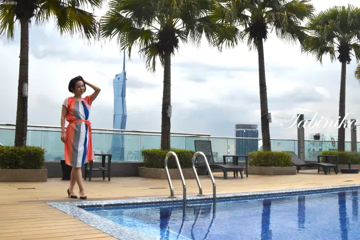 Woman in colorful dress at rooftop pool with KL skyline view, featuring the iconic Menara Maxis tower.