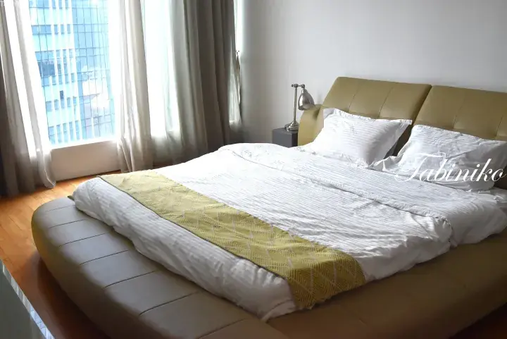 Bright hotel bedroom with a double bed, white linens, and a yellow throw, featuring large windows with city views.