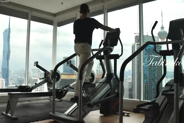 Man exercising on a stair climber in a high-rise gym overlooking the Kuala Lumpur skyline, including the Merdeka 118 tower.