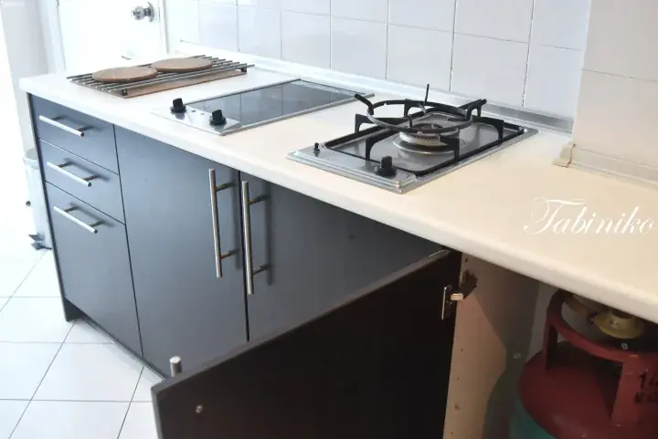 Modern kitchen with grey cabinets, white countertop, gas stove, and red LPG tank visible under the counter.