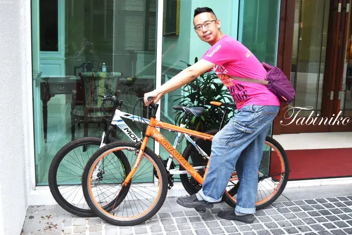 A man in a pink shirt stands with two bicycles, an orange Gainway and a white one, outside a building with large glass windows.