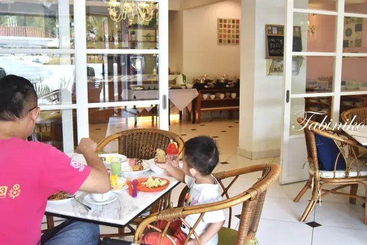 Father and son enjoying breakfast at a hotel dining area with buffet setup and rattan furniture.
