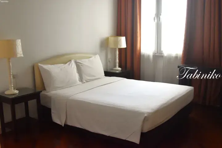 A neatly made double bed with white linens in a hotel room, featuring bedside tables, lamps, and brown curtains.