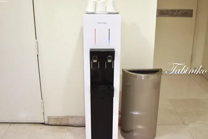 Coway water dispenser with hot and cold buttons next to a metal trash bin in a building hallway.