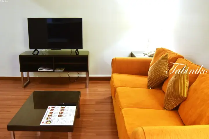 Bright living room with an orange sofa, flat-screen TV, and coffee table menu at a serviced residence.