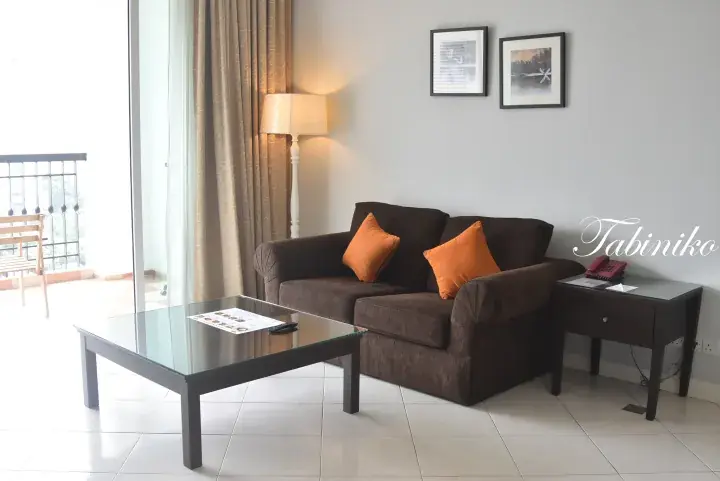 Modern apartment living room with brown sofa, orange cushions, glass coffee table, and balcony access.