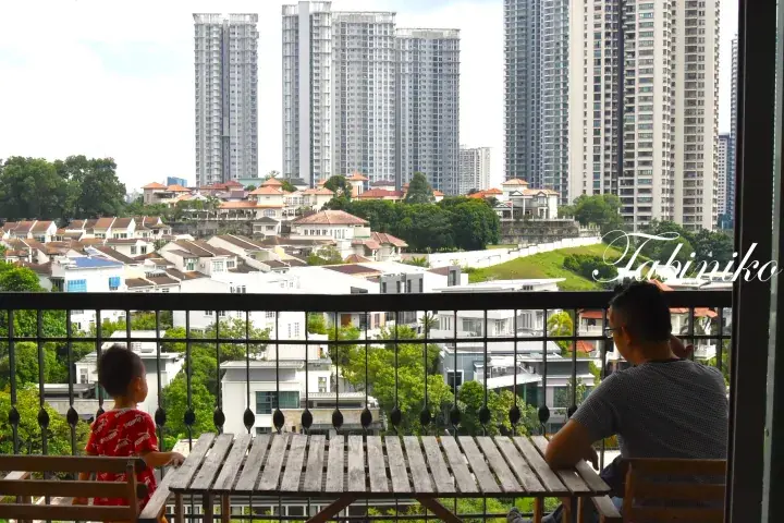 A man and child on a balcony overlooking a mix of landed homes and high-rise apartments in a Malaysian urban area.