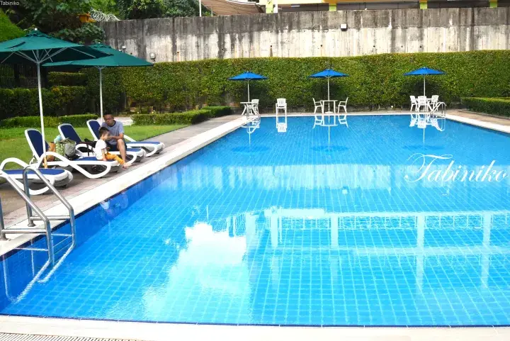 A family relaxing by a bright blue swimming pool with lounge chairs and umbrellas at a Malaysia property.