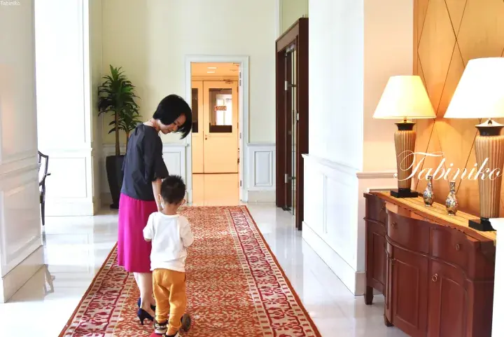 Woman and child walking on a patterned hallway carpet in a hotel corridor with wooden furniture.