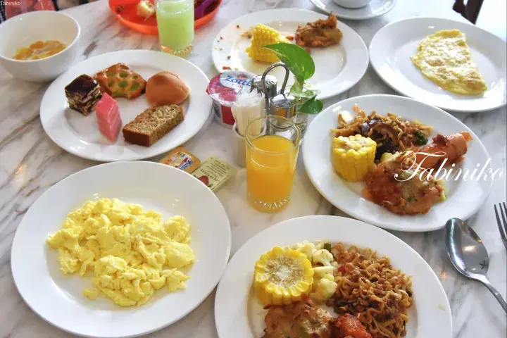 A breakfast spread featuring scrambled eggs, omelet, corn, noodles, and pastries on a marble table.