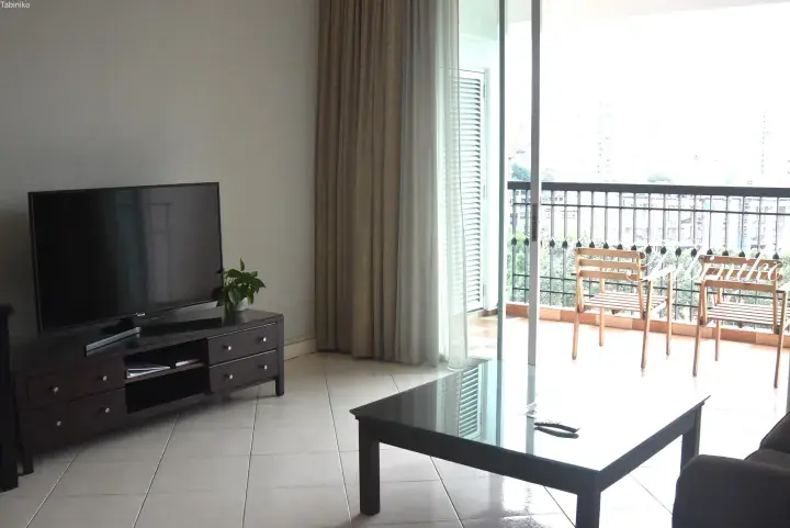 Modern living room with flat-screen TV, glass coffee table, and balcony with wooden chairs overlooking city view