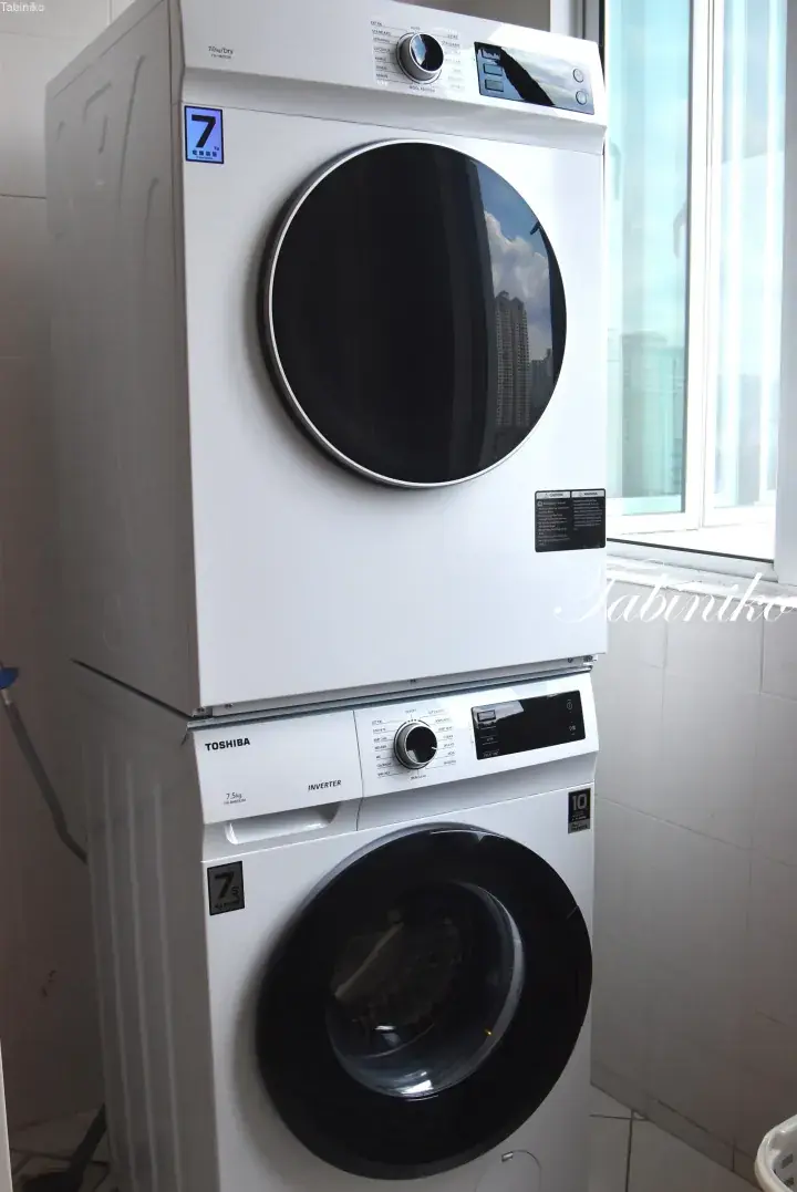 Stacked white Toshiba washer and dryer unit in a modern apartment laundry room with a window view.