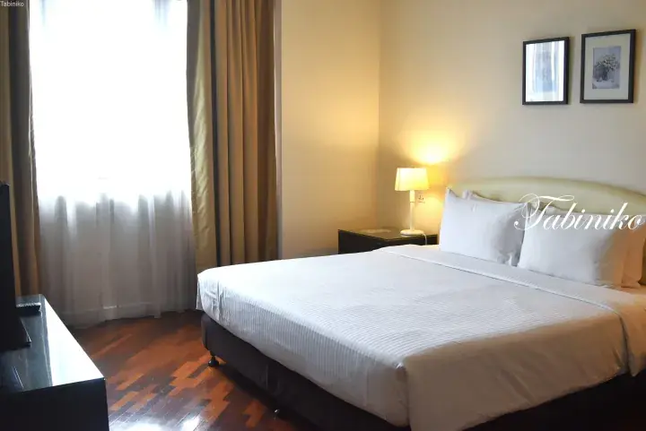 Spacious hotel bedroom with a double bed, wooden flooring, and a window with sheer curtains.