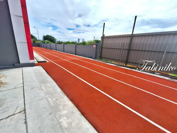 Red running track with white lane lines alongside a grey wall and fence under a partly cloudy sky.
