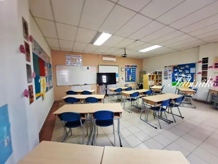 Bright classroom with wooden desks, blue chairs, whiteboard, and educational posters on the walls.