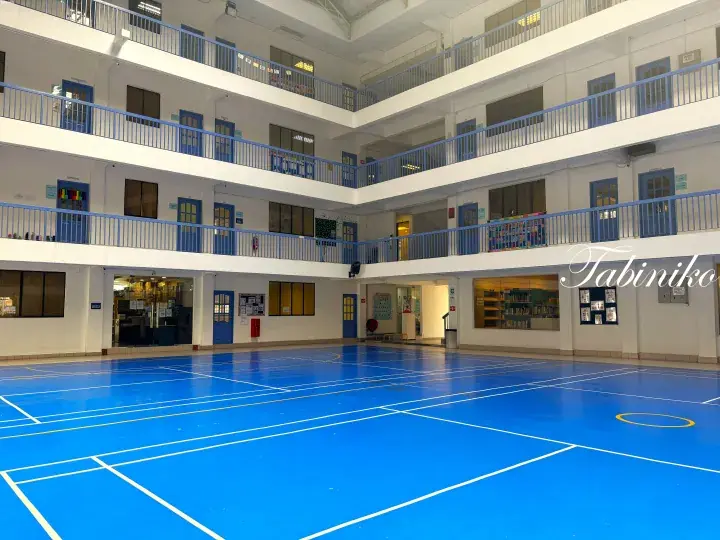 Spacious indoor sports hall with blue flooring and white walls, surrounded by multiple levels of classrooms with blue railings.