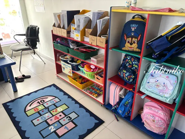 Colorful classroom corner with toy shelves, children&rsquo;s backpacks, and a hopscotch rug.