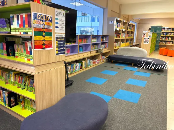 Colorful children&rsquo;s library with wooden bookshelves, blue carpet squares, and a grey reading bench.