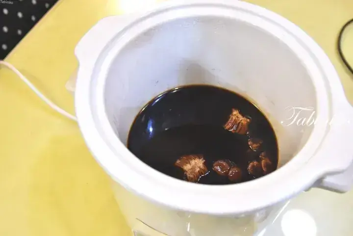 Close-up of a white slow cooker pot containing dark soy sauce and dried shiitake mushrooms.