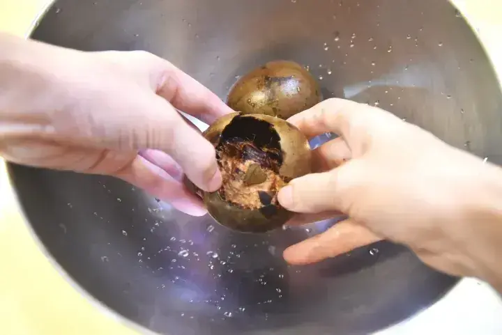 Hands peeling open a brown, round fruit over a metal bowl, revealing its fibrous interior.