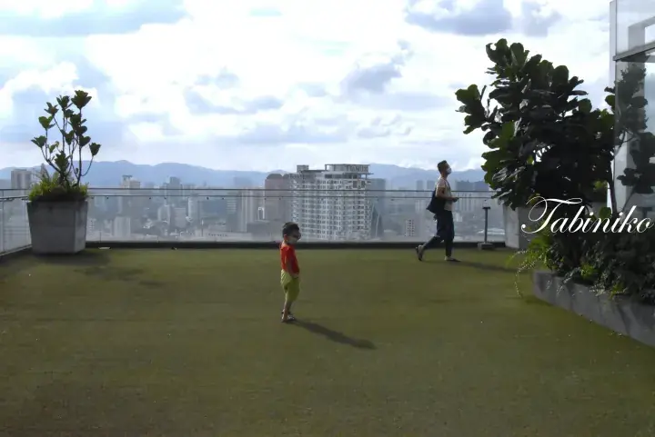 Child and adult enjoying a rooftop garden with artificial turf and city skyline views in Malaysia.