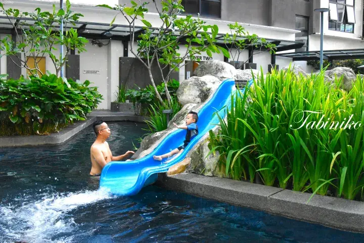 Father and child enjoying a blue water slide at a resort pool surrounded by lush greenery.