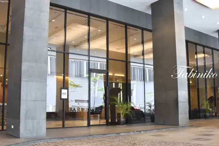 Modern hotel entrance with large glass windows, grey stone pillars, and a visible lobby interior.