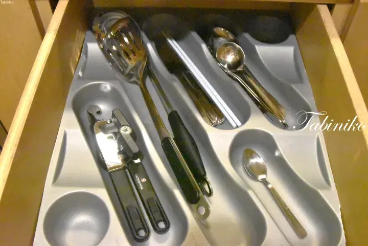 Organized kitchen drawer with a can opener, slotted spoon, tongs, and spoons neatly arranged in a grey tray.