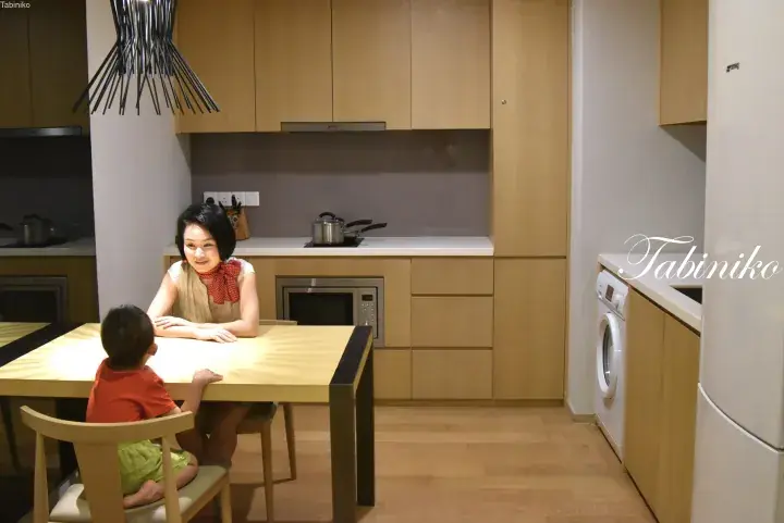 Mother and child sitting at a wooden dining table in a modern kitchen with light wood cabinetry and a washing machine.