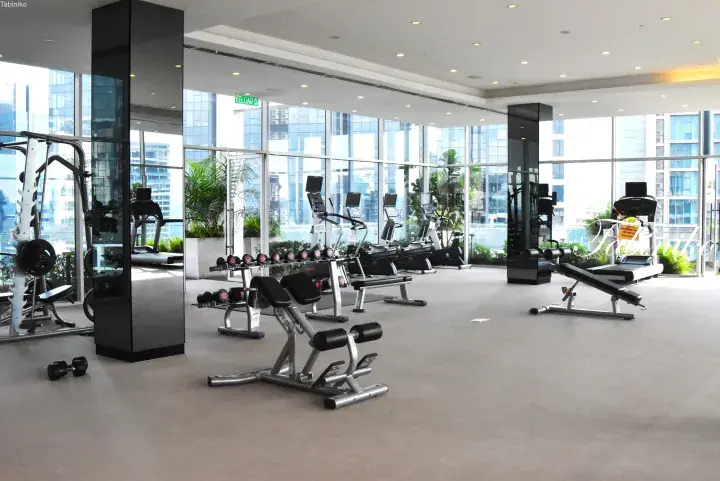 Modern gym with weight benches, dumbbells, and cardio machines overlooking city skyline through large windows.