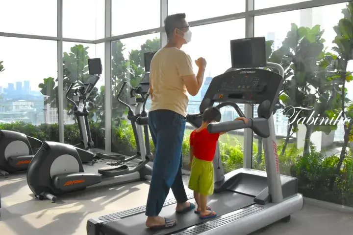 A man and young child walking on a treadmill in a gym with large windows overlooking a city skyline.