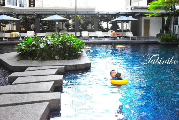 A father and child enjoy the swimming pool at a modern residential complex with landscaped greenery and lounge chairs.