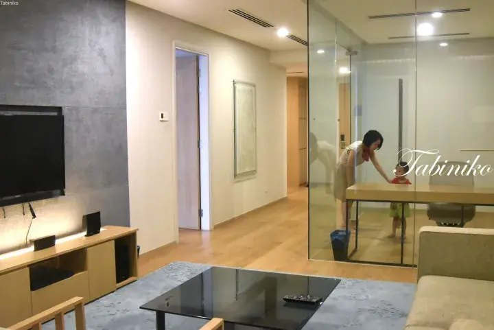 Modern apartment living room with wooden floors, TV, and glass-walled dining area where a woman and child are present.
