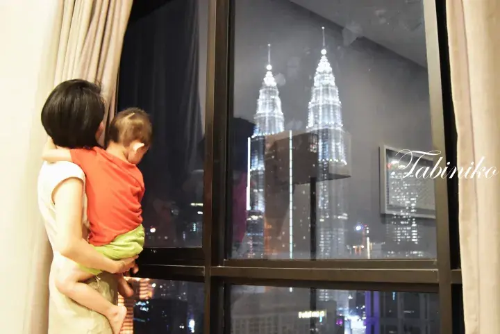 Mother holding child looking out window at illuminated Petronas Twin Towers in Kuala Lumpur.