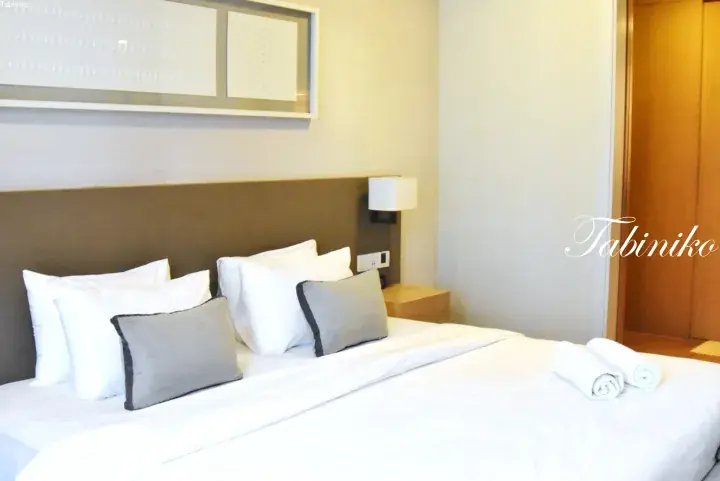 Modern hotel bedroom with a neatly made bed, white linens, grey accent pillows, and rolled towels.