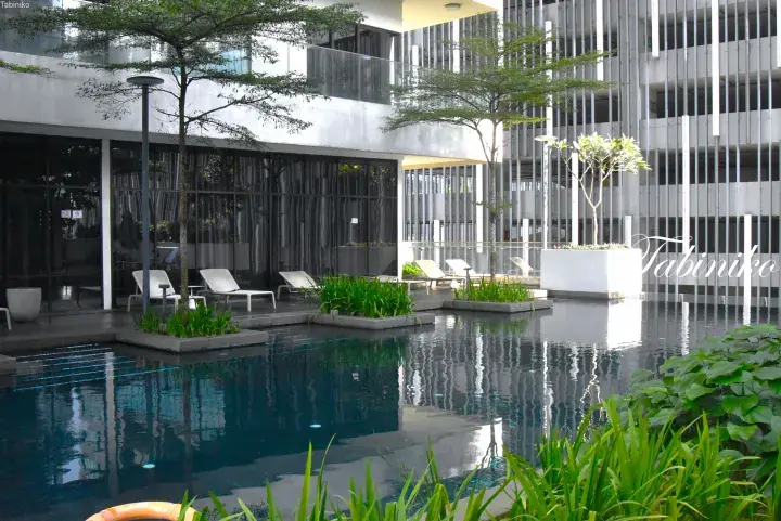 Modern resort pool with white lounge chairs, lush greenery, and reflective water surrounded by contemporary architecture.
