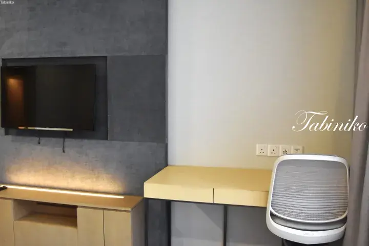 Modern hotel room featuring a wall-mounted TV, a light wood desk with a mesh office chair, and a grey textured wall.